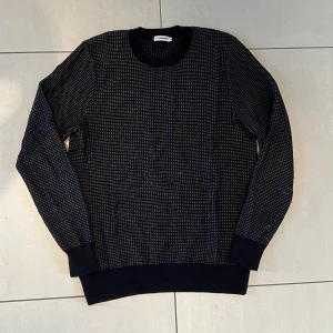 Svart stickad tröja J.Lindeberg M - Snygg svart stickad tröja från J.Lindeberg i storlek M. Tröjan har ett diskret rutigt mönster i vitt och ribbade muddar vid ärmslut, hals och nederkant. Perfekt för dig som gillar stilrena och klassiska plagg med en twist. Lite nopprig men går att fixa!!🤩🤩