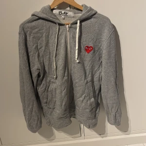 Grå hoodie Comme des Garçons Play - Grå hoodie med dragkedja från Comme des Garçons Play. Tröjan har huva med vita snören och röda hjärtan med ögon både fram och bak. Stora fickor framtill och PLAY-logga på ryggen. Köpt för: 2800 kr. Mitt pris: 900kr. Tveka inte på att höra av er vid mer frågor. 