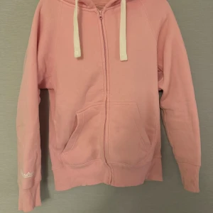 Rosa hoodie från russemerch med dragkedja - Mysig ljusrosa hoodie från Russell med vit snörning och hel dragkedja framtill. Perfekt för chill dagar och enkel att matcha med jeans eller mjukisbyxor.