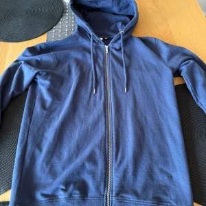 Marinblå ziphoodie från Minimum - Säljer en marinblå hoodie från Minimum med dragkedja och snörning i huvan. Tröjan är i mjuk bomull, har långa ärmar och ribbade muddar. Perfekt för chill dagar och enkel att matcha med jeans eller joggers. Snygg och enkel design utan mönster.