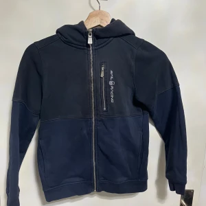 Marinblå & svart kofta från Sail Racing - Snygg marinblå halv och halv svart kofta från Sail Racing med huva och hel dragkedja framtill. Tröjan har en bröstficka med dragkedja och logotyp, samt långa ärmar och ribbade muddar. Perfekt för dig som gillar sportig och avslappnad stil. Storlek 140 cm