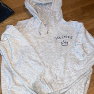Ljusgrå hoodie ma chérie från Gina tricot  - Säljer en ljusgrå hoodie med huva och snörning. På bröstet finns ett coolt MA CHÉRIE-tryck och en krona i mörkgrått. Tröjan har långa ärmar och ribbade muddar. Perfekt för chill dagar och enkel att matcha med jeans eller joggers. Endast använt den en gång då den va för stor för mig ❤️ 