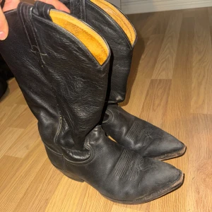 Svarta cowboy boots i skinn - Säljer ett par klassiska svarta cowboy boots i äkta skinn med spetsig tå och låg klack. Bootsens insida är ljust brun och de har snygga broderade detaljer framtill