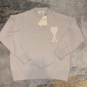 Ljusbeige stickad tröja från Ami - Säljer en ljusbeige stickad tröja från Ami med rund hals och ribbade muddar. Tröjan har Ami:s ikoniska hjärta och A-logga på bröstet. Perfekt för dig som gillar stilrena och trendiga plagg med en twist.