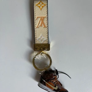 Vuitton x Nike nyckelring sneaker - Exklusiv Vuitton x Nike nyckelring med en miniatyr sneaker i brunt, orange och guld. Remmen är beige med LV-logga i gult och vitt, samt guldfärgad metallring. Snygg accessoar med ikoniska monogramdetaljer och cool design.