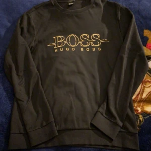 Svart Hugo Boss sweatshirt slim fit - Svart sweatshirt från Hugo Boss med stort BOSS-tryck i guld på bröstet. Slim fit-modell med rund hals och långa ärmar. Mjuk bomullskvalitet och ribbade muddar vid ärmslut och nederkant. Perfekt för dig som gillar stilrena och klassiska märkesplagg.