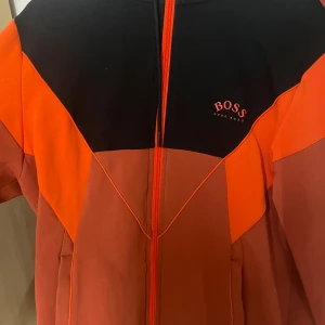 Svart och orange vindjacka från Hugo Boss - Snygg vindjacka från Hugo Boss i svart och orange med coola geometriska paneler. Jackan har hel dragkedja framtill, huva med snörning och logga på bröstet. Tillverkad i mjuk bomull och polyesterblandning, perfekt för dig som gillar sportig och modern stil.