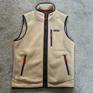 Beige fleeceväst från Patagonia - Säljer en beige fleeceväst från Patagonia med marinblå detaljer och röda dragkedjor. Västen har hög krage, tre fickor med dragkedja och klassisk Patagonia-logga på bröstet. Perfekt för lager-på-lager och outdoor-vibe.