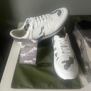 Valentino Garavani sneakers camo - Snygga Valentino Garavani sneakers i vitt med grå och beige camouflage-mönster. Klassisk rund tå, platt sula och snörning. Märkeslogga på plösen och insidan. Tillverkade i skinn och syntet för en lyxig känsla. Perfekta för dig som vill sticka ut med stil.