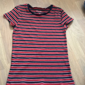 Randig topp från Etirel XS - Säljer en randig topp från Etirel i storlek XS. T-shirten har röda, marinblå och vita horisontella ränder, rund halsringning och korta ärmar. Materialet är  stretchigt, vilket gör den bekväm och enkel att bära.