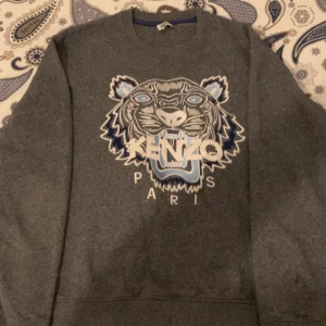 Grå Kenzo sweatshirt med tigertryck - Säljer en grå sweatshirt från Kenzo med det ikoniska tigerhuvudet broderat i blått, vitt och ljusblått på bröstet. Tröjan har rund hals och lång ärm, perfekt för dig som gillar streetstyle och vill sticka ut med ett coolt print.