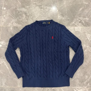 Marinblå kabelstickad tröja Polo Ralph Lauren - Snygg marinblå kabelstickad tröja från Polo Ralph Lauren med klassisk röd broderad logga på bröstet. Tröjan har rund hals, långa ärmar och ribbade muddar. Perfekt för dig som gillar stilren och tidlös design.