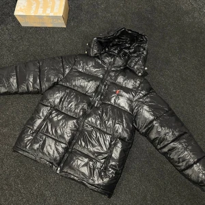 Svart pufferjacka från Polo Ralph Lauren - Svart pufferjacka från Polo Ralph Lauren med glansig finish och röd logga på bröstet. Jackan har huva, två stora fickor med dragkedja och en kraftig frontdragkedja. Perfekt för kalla dagar och har en riktigt schysst streetstil.