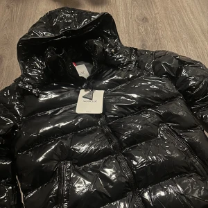 Svart Moncler pufferjacka med huva - Säljer en svart, glansig pufferjacka från Moncler med huva och dragkedja framtill. Jackan har quiltad design, två sidofickor med dragkedja och en cool Moncler-logga på ärmen. Perfekt för dig som vill ha en snygg och varm jacka med streetkänsla.