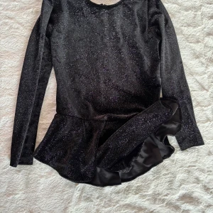 Svart glittrig konståkningklänning med volang - Supersnygg svart klänning med glittrigt tyg och långa ärmar. Modellen är kort med en volangkant nedtill och har en underbyxa. Materialet känns mjukt och stretchigt, perfekt för att sticka ut med lite extra glitter.