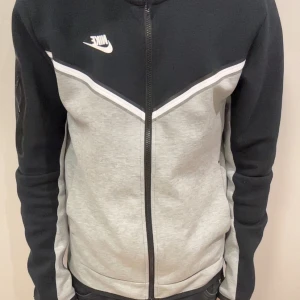 Nike svart och grå tech fleece  - Nike tech fleece i svart och ljusgrått med vit logga på bröstet. Koftan har hel dragkedja framtill, långärmade ärmar och en snygg detalj med dragkedjeficka på ena ärmen. Materialet är mjukt och känns sportigt, perfekt för chill eller träning. Pris ej hugget i sten så det är bara att höra av sig.🤩