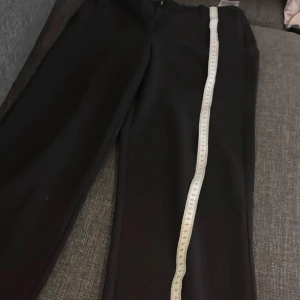 Kostymbyxor  - Snygga svarta kostymbyxor med raka ben och hög midja. Byxorna har bälteshällor, fickor och stängs med dragkedja och knapp. Perfekta för en clean och stilren look. Materialet känns mjukt och har ett fint fall.