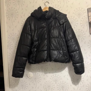 Svart pufferjacka från NA-KD - Säljer en svart pufferjacka från NA-KD med snygg croppad passform och huva. Jackan har quiltad design, dragkedja framtill och två fickor med knappstängning. Materialet är syntet med skinnlook som ger en cool vibe. Perfekt för kalla dagar när du vill se extra fräsch ut.