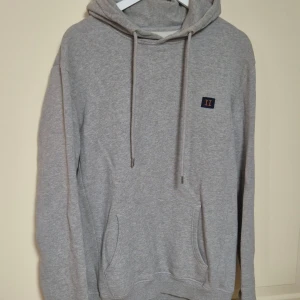 Grå hoodie från Les Deux med snörning L - Säljer en stilren grå hoodie från Les Deux med snörning i huvan och en liten broderad detalj på bröstet. Tröjan har en klassisk känguruficka och långa ärmar, perfekt för chill dagar. Mjuk bomullskänsla och relaxed passform. I mycket fint skick!