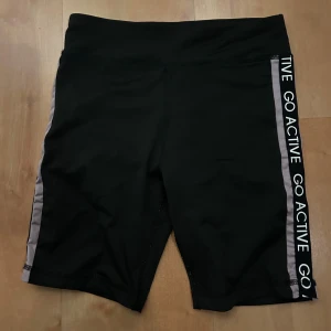 Tajta svarta träningsshorts KappAhl - Svarta, medellånga till korta cykelshorts från KappAhl med bred resår i midjan och texten 'GO ACTIVE' längs sidorna på ett band i lila. Tajt passform och stretchigt material som passar perfekt till träning.