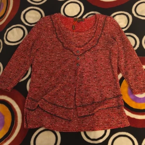 Röd stickad kofta från Naveed - Säljer en röd stickad kofta från Naveed i storlek M. Koftan har tre dekorativa knappar framtill, ett snyggt lager-på-lager utseende och subtila svarta och vita inslag i garnet. Den har trekvartsärm och en mjuk, rundad halsringning med diskret kant.