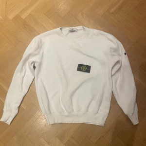 Vit Stone Island sweatshirt med patch - Vit sweatshirt från Stone Island med klassisk rund hals och långärmad passform. Tröjan har den ikoniska Stone Island-patchen på vänster ärm, fästad med svarta knappar. Använd men utan större defekter. 
