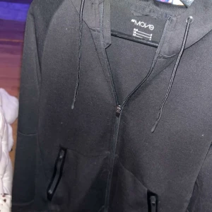 Svart hoodie från H&M Move med dragkedja - Svart hoodie från H&M Move med hel dragkedja, snörning i huvan och två fickor framtill. Perfekt för chill eller träning, med normal passform och mjukt material som känns skönt mot huden.