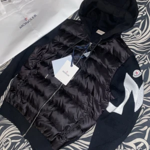 Svart pufferjacka med huva från Moncler - Snygg svart pufferjacka från Moncler med huva och dragkedja. Jackan har quiltad framsida i glansigt material, stickade ärmar med vit detalj och Moncler-logga på ärmen. Perfekt för dig som vill ha en stilren och exklusiv look.