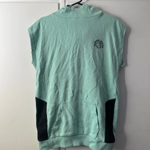 Mintgrön ärmlös hoodie från F&F - Snygg mintgrön ärmlös hoodie från F&F med svart mesh-detalj på sidorna och magficka. Tröjan har huva och tryckt logga på bröstet. Perfekt för en chill och sportig look. Tillverkad i mjuk bomullsmix.