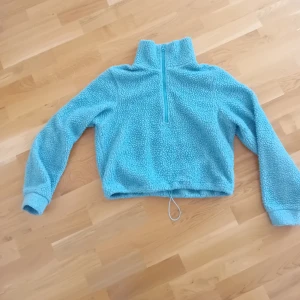 Blå fleece-jacka från Bik Bok XS - Mysig ljusblå fleece-tröja från Bik Bok i storlek XS. Tröjan har en hög krage, half zip-dragkedja framtill och långa ärmar. Croppad passform med snörning i nederkant för extra stilpoäng. Perfekt för kyliga dagar när du vill vara både bekväm och snygg.