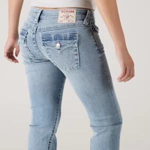 True Religion ljusblå bootcut jeans - Säljer ett par ljusblå jeans från True Religion med bootcut passform och låg midja. Jeansen har snygga detaljerade bakfickor med lock och silverfärgade knappar samt klassisk logopatch bak i midjan. Perfekta för dig som gillar en avslappnad men trendig look.
