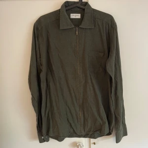 Olivgrön overshirt från John Henric - Olivgrön overshirt från John Henric med dragkedja framtill, klassisk krage och bröstficka. Långärmad modell i mjukt bomullstyg, perfekt att slänga över en t-shirt. Enkel och stilren design som funkar till många outfits. Nypris 899kr