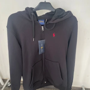 Svart hoodie från Polo Ralph Lauren - Svart hoodie från Polo Ralph Lauren med dragkedja, huva och snörning. Klassisk röd broderad logga på bröstet och stora fickor framtill. Tillverkad i mjuk bomullsmix, perfekt för chill dagar eller när du vill ha en clean look.storlek L men sitter mer som en M.