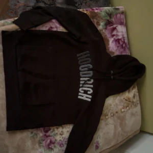 Svart HOODRICH hoodie med tryck - Svart hoodie från HOODRICH med stort vitt tryck längs bröstet. Klassisk modell med huva och snörning samt en stor magficka. Perfekt för dig som gillar streetwear och vill ha en skön och stilren tröja.