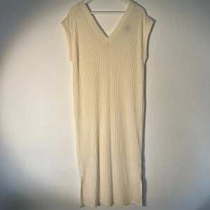 Ribbad beige långklänning XS - Ribbad beige långklänning med v-ringning och ärmlös design. Klänningen har en rak passform och slits nertill för extra rörelsefrihet. Perfekt för en stilren och avslappnad look. Ny med prislapp!