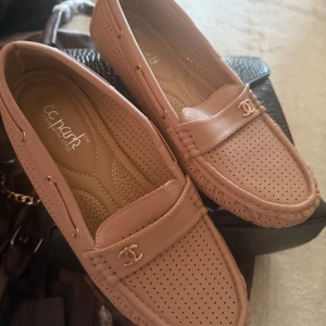 Beige loafers med gulddetalj - Snygga beige loafers i skinn med perforerat mönster och guldfärgad detalj framtill. Skorna har klassisk rund tå och platt sula, perfekta för en stilren look. Lätta att matcha till olika outfits och riktigt bekväma att bära hela dagen.