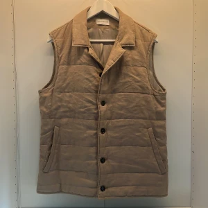 Beige quiltad dunväst These Glory Days - Snygg beige quiltad dunväst från These Glory Days i storlek medium. Västen har klassisk krage, knäppning med svarta knappar och två fickor framtill. Perfekt lager-på-lager-plagg med stilren look och mjukt material.