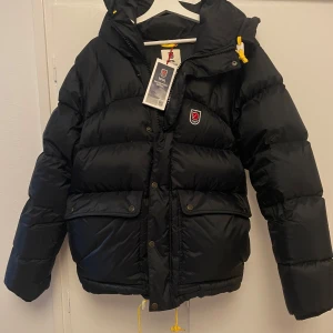Vinterjacka från Fjällräven  - Snygg svart dunjacka från Fjällräven med huva och gula detaljer. Jackan är varm, har dragkedja och knappar framtill samt två stora fickor. Perfekt för kalla dagar och har klassisk Fjällräven-logga på bröstet. Helt ny med tagg kvar.