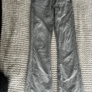 Grå bootcut jeans med broderade fickor - Snygga grå jeans med bootcut passform och unika broderade detaljer på bakfickorna. Jeansen har en lätt tvättad look och är tillverkade i klassiskt denimtyg. Perfekta för dig som vill sticka ut med coola detaljer och en avslappnad vibe. Saknas knappar på båda fickorna där bakom.