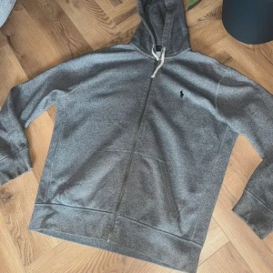 Grå hoodie från Polo Ralph Lauren - Hoodie Polo Ralph Lauren med dragkedja och huva med vita snören. Klassisk logga  på bröstet.