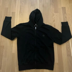 Svart Moncler hoodie med dragkedja - Svart hoodie från Moncler med hel dragkedja framtill, huva med snörning och klassisk Moncler-logga på bröstet. Tröjan har två stora fickor framtill och är tillverkad i mjuk bomull. Perfekt för en chill och stilren look.