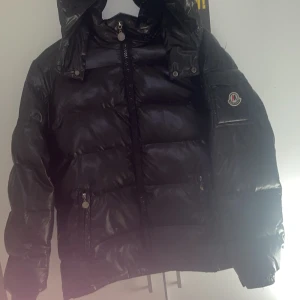 Svart pufferjacka från M Fashion - Snygg svart pufferjacka från M Fashion med glansig finish, dragkedja framtill och praktiska fickor. Jackan har huva, snörning i nederkant och en patch med logga på ärmen. Perfekt för kalla dagar när du vill hålla stilen. Vet tyvär inte storlek men den är lite för stor för mig och jag är 173
