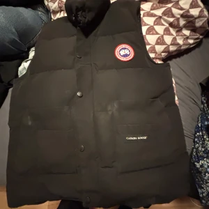 Svart dunväst från Canada Goose - Svart dunväst från Canada Goose med klassisk logga på bröstet och stora fickor framtill. Västen har hög krage, tryckknappar.  Finns några fläckar bara ta en trasa med vatten och gnugga så är dem borta  