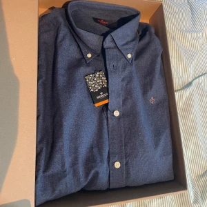 Blå skjorta från Morris Stockholm - Snygg blå skjorta från Morris Stockholm med klassisk button down-krage och vita knappar. Skjortan har en broderad guldfärgad lilja på bröstet och är tillverkad i mjuk bomull. Perfekt för dig som gillar stilrena och tidlösa plagg. Prislapp sitter kvar ock skickas i original förpackning. Nypris 1499kr