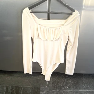 Vit långärmad bodysuit med rynk - Säljer en stilren vit bodysuit med långa ärmar och fyrkantig urringning. Toppen har en snygg rynkdetalj framtill och är tillverkad i ett mjukt, stretchigt material. Perfekt att styla med jeans eller kjol för en clean look.