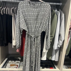 Grå långklänning - Säljer en grå långklänning med vitt blommigt mönster. Klänningen har långa ärmar, rund halsringning och ett avtagbart knytband i midjan som ger en snygg siluett. Perfekt för dig som gillar en avslappnad men ändå stilren look. Strl S/M