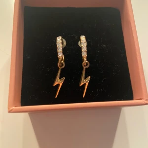 Guldiga blixtörhängen med stenar - Snygga örhängen i guld med blixtformade hängen och små glittriga stenar på ringen. Perfekt för dig som vill sticka ut med coola detaljer.