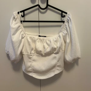 Vit croppad blus från Gina Tricot - Superfin vit blus från Gina Tricot med puffiga korta ärmar och offshoulder-modell. Toppen är croppad och har en lätt strukturerad yta som ger en cool känsla. Perfekt för dig som gillar trendiga och feminina plagg med snygg passform.