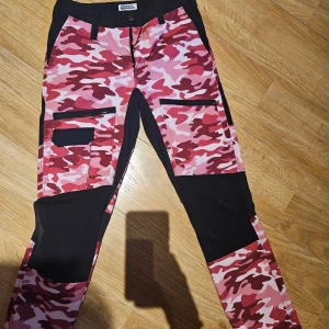 Camouflage byxa i rosa - Camouflagebyxa inköpt på narurkompaniet / fjällräven . STRL M . Använda sparsamt,  i gott skick. Två större fickor med dragkedja fram plus liten på en benet med kardborreband. En fika med dragkedja bak.  Tyvärr för små numera åt mig . 