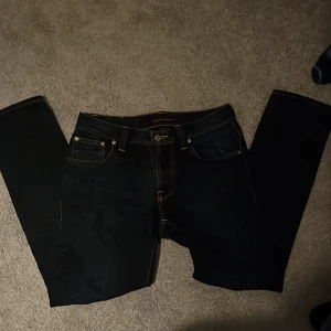 Mörkblå Nudie Jeans - Säljer ett par mörkblåa jeans från Nudie Jeans med klassiska kontrastsömmar och snygga bakfickor med vågig brodyr. Jeansen har fem fickor, normal passform och är tillverkade i slitstarkt bomullstyg. Perfekta för dig som gillar stilrena och tidlösa jeans.